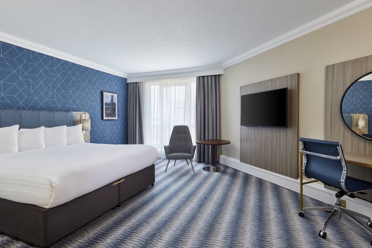Une chambre d’hôtel avec un grand lit, un bureau, une chaise, une télévision et une fenêtre avec des rideaux.