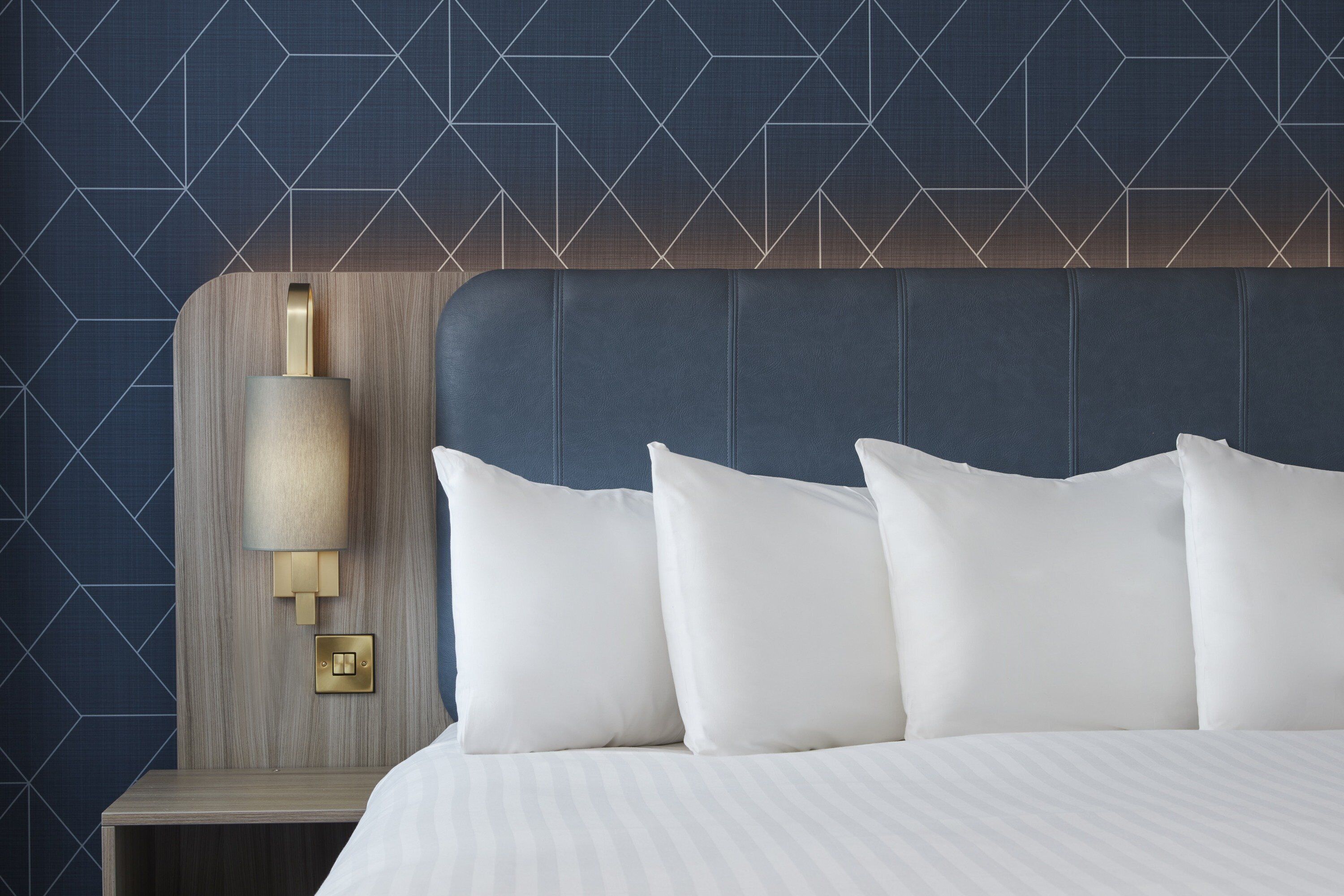 A hotel room with a bed, white pillows, a headboard, and a wall-mounted nightstand with a lamp.