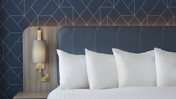 A hotel room with a bed, white pillows, a headboard, and a wall-mounted nightstand with a lamp.
