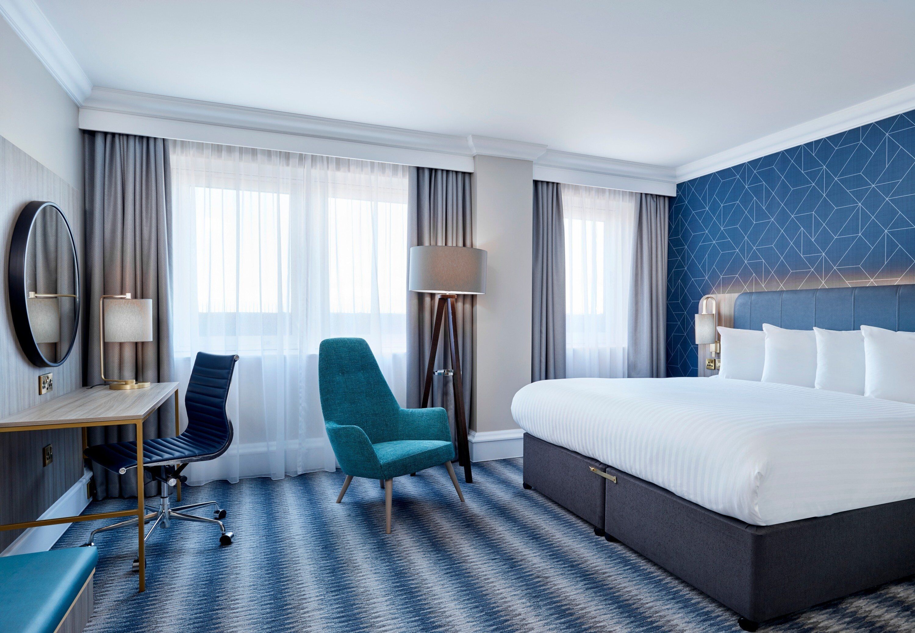 A hotel room with a large bed, a desk with a chair, a standing lamp, and a large mirror.