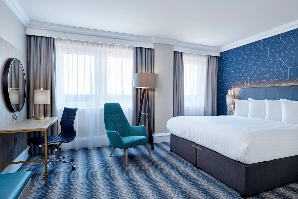Une chambre d’hôtel avec un grand lit, un bureau avec une chaise, une lampe sur pied et un grand miroir.