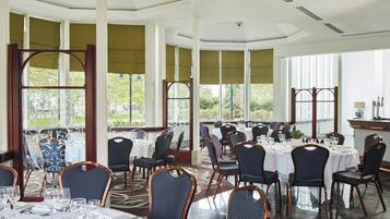 A restaurant with round tables and chairs, large windows, and a view of greenery outside.