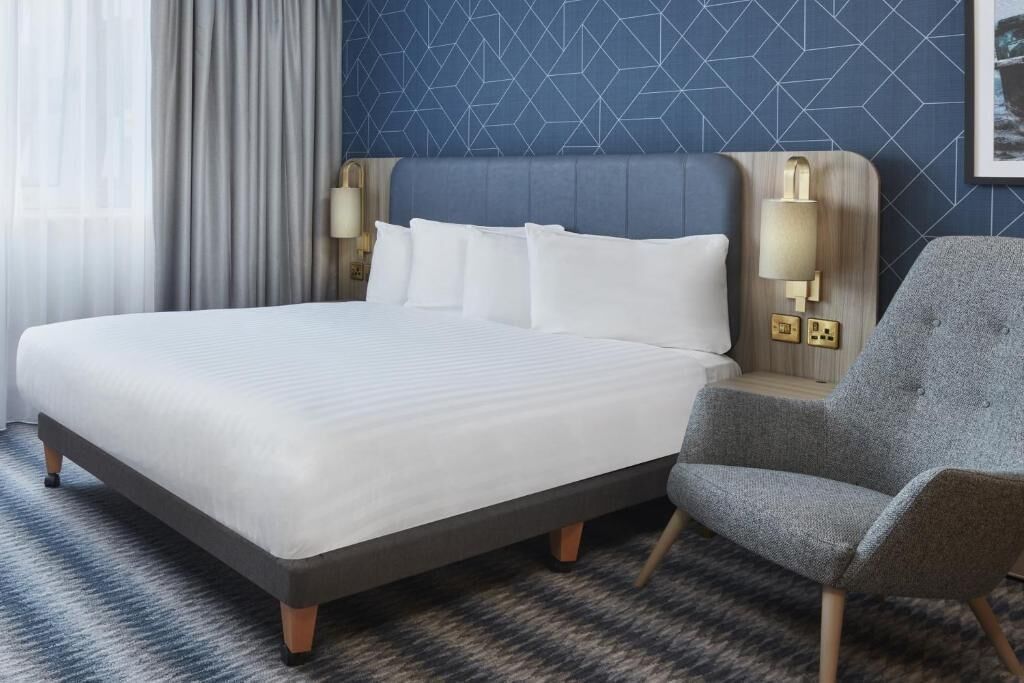 A hotel room with a large bed, two bedside lamps, a grey armchair, and a patterned wall.