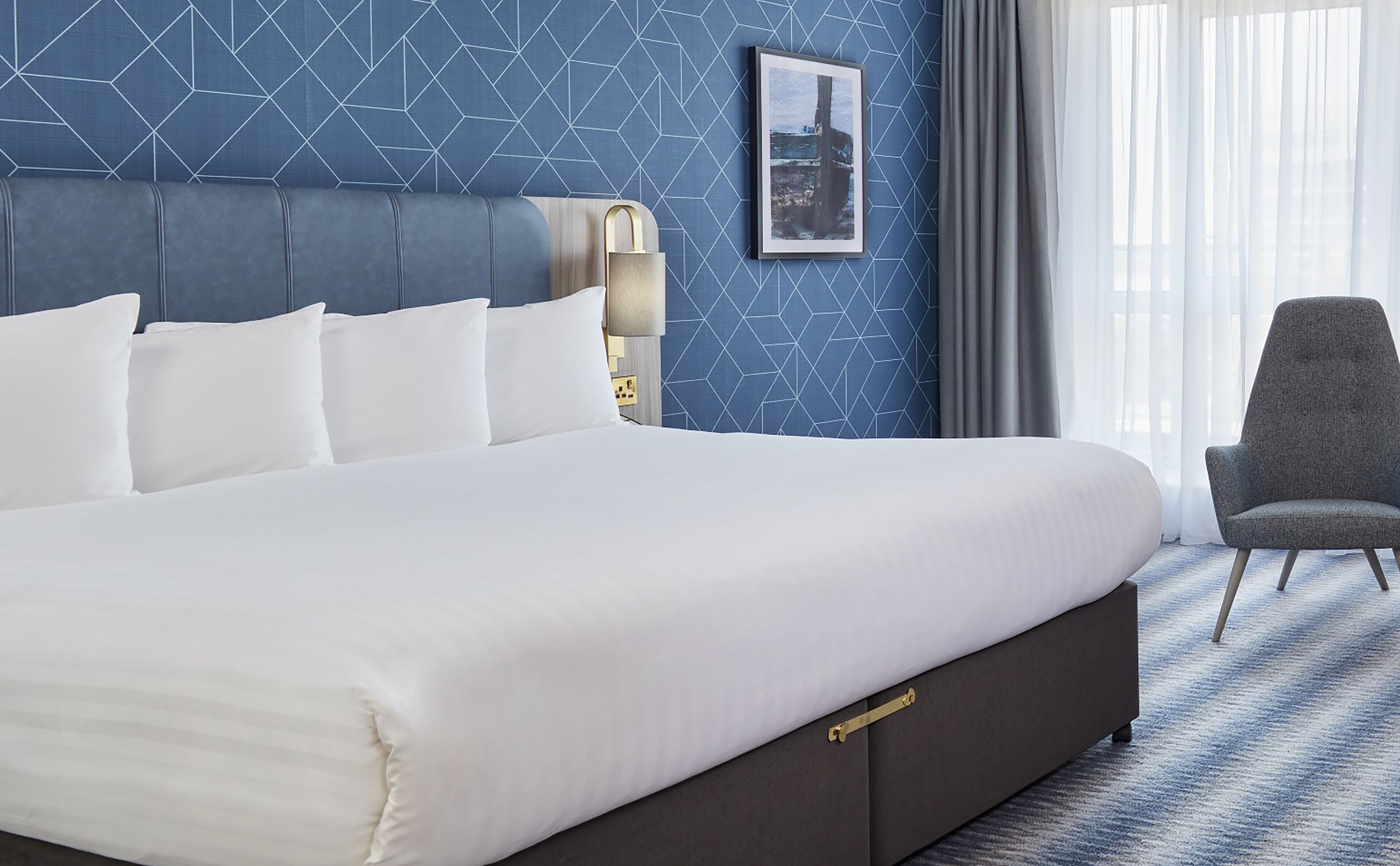 Une chambre d’hôtel avec un grand lit, une tête de lit, un tableau encadré au mur et une chaise.
