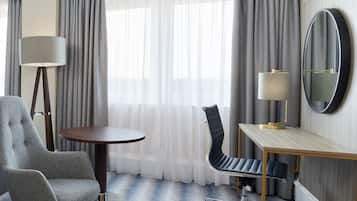 A hotel room with a grey upholstered armchair, a round table, a desk with a chair, a floor lamp, and a mirror.