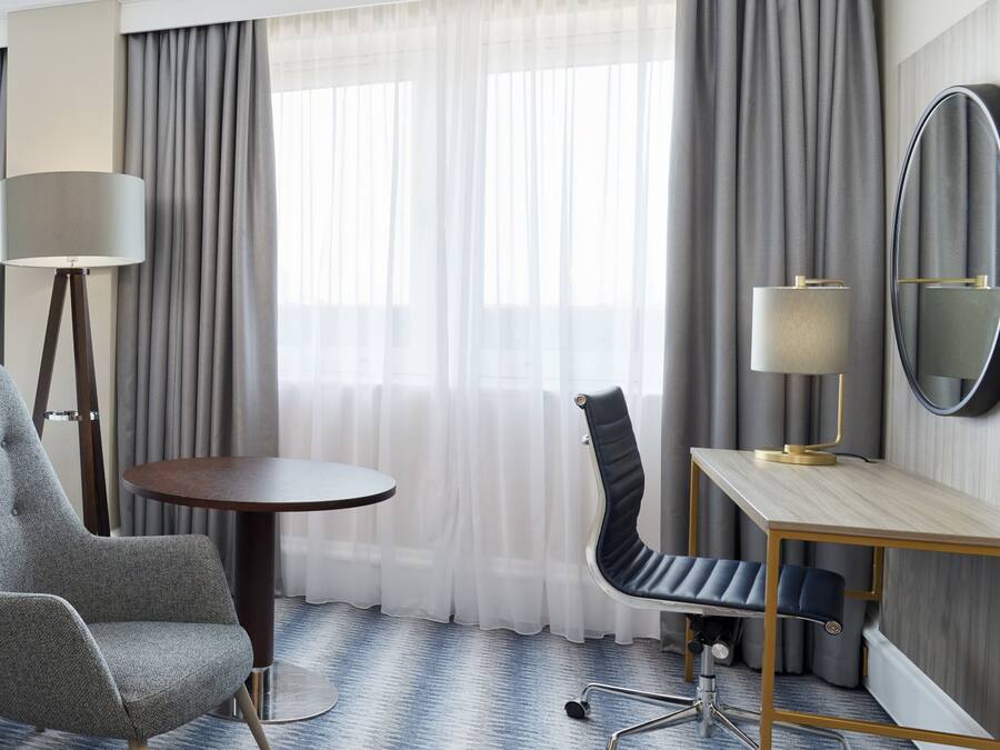A hotel room with a grey upholstered armchair, a round table, a desk with a chair, a floor lamp, and a mirror.
