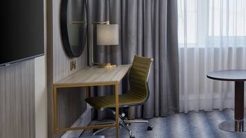 A hotel room with a wooden desk, a green upholstered chair, a round table, a TV, and a lamp.