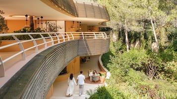 Um edifício moderno com varanda curva e área de estar ao ar livre, rodeado por vegetação.