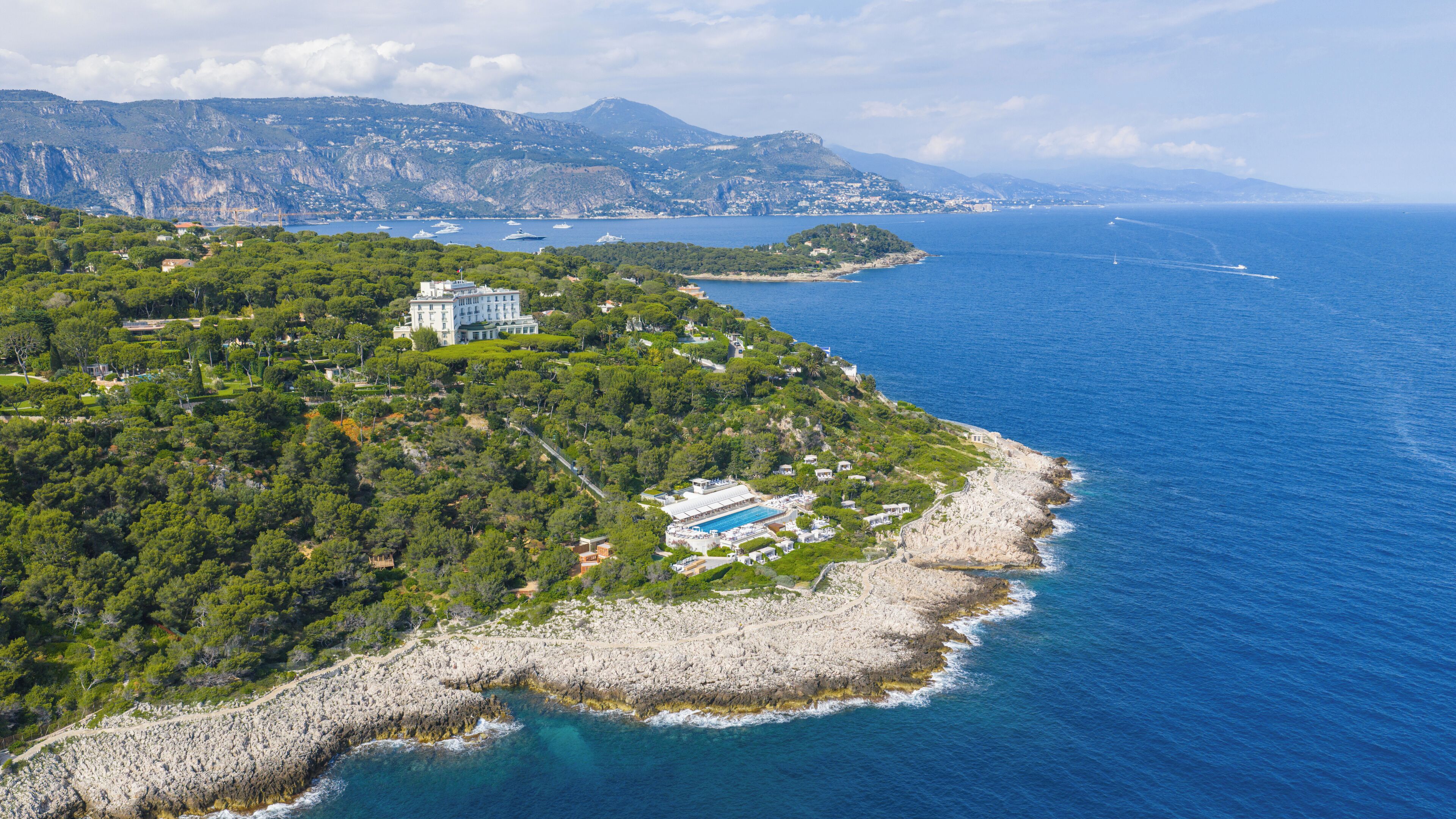Vue aérienne d’une propriété située sur la côte, avec un grand bâtiment, une piscine et une plage rocheuse.