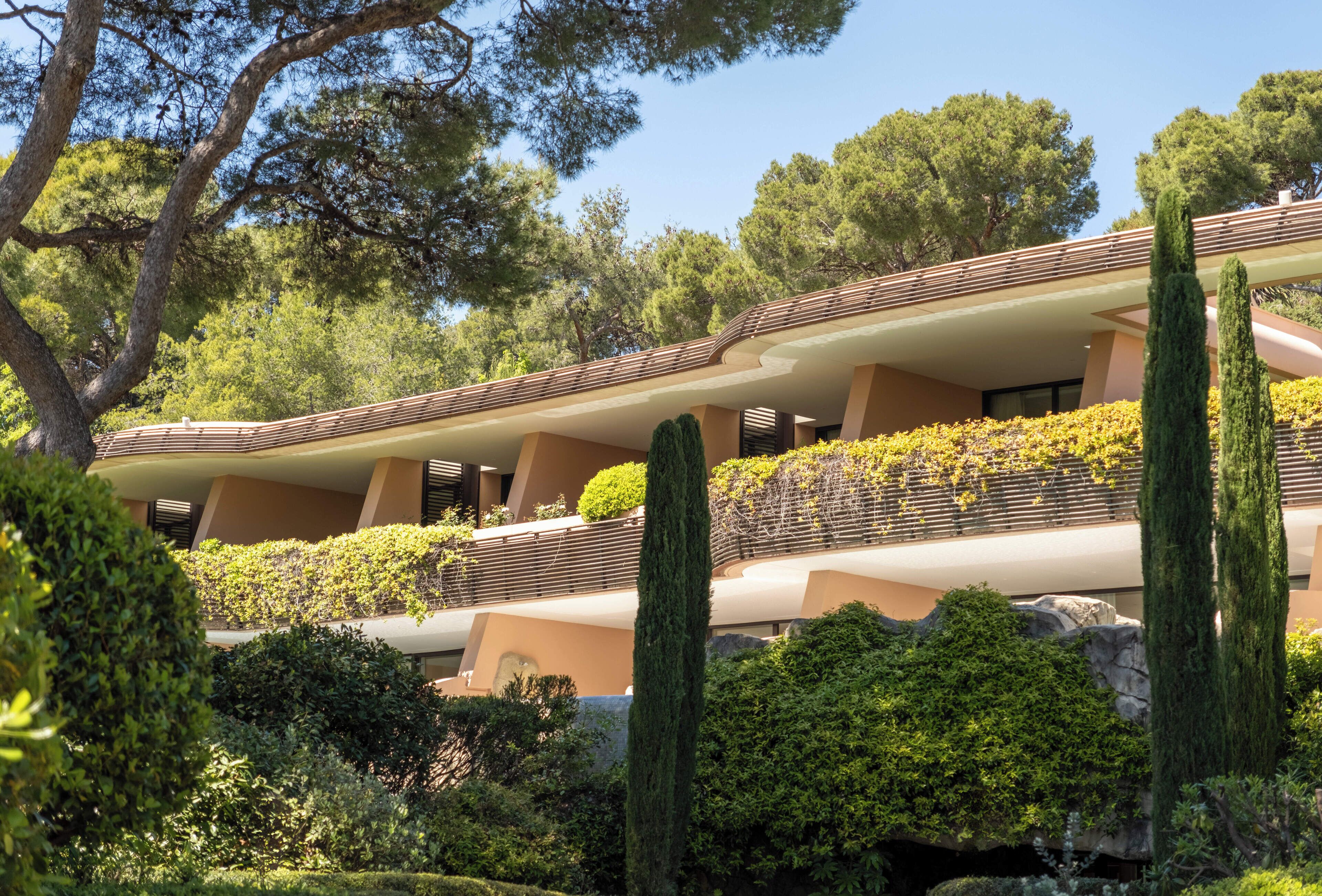 Un edificio moderno con varios balcones, rodeado de abundante vegetación y altos cipreses.