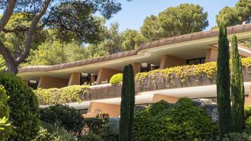 Un edificio moderno con varios balcones, rodeado de abundante vegetación y altos cipreses.