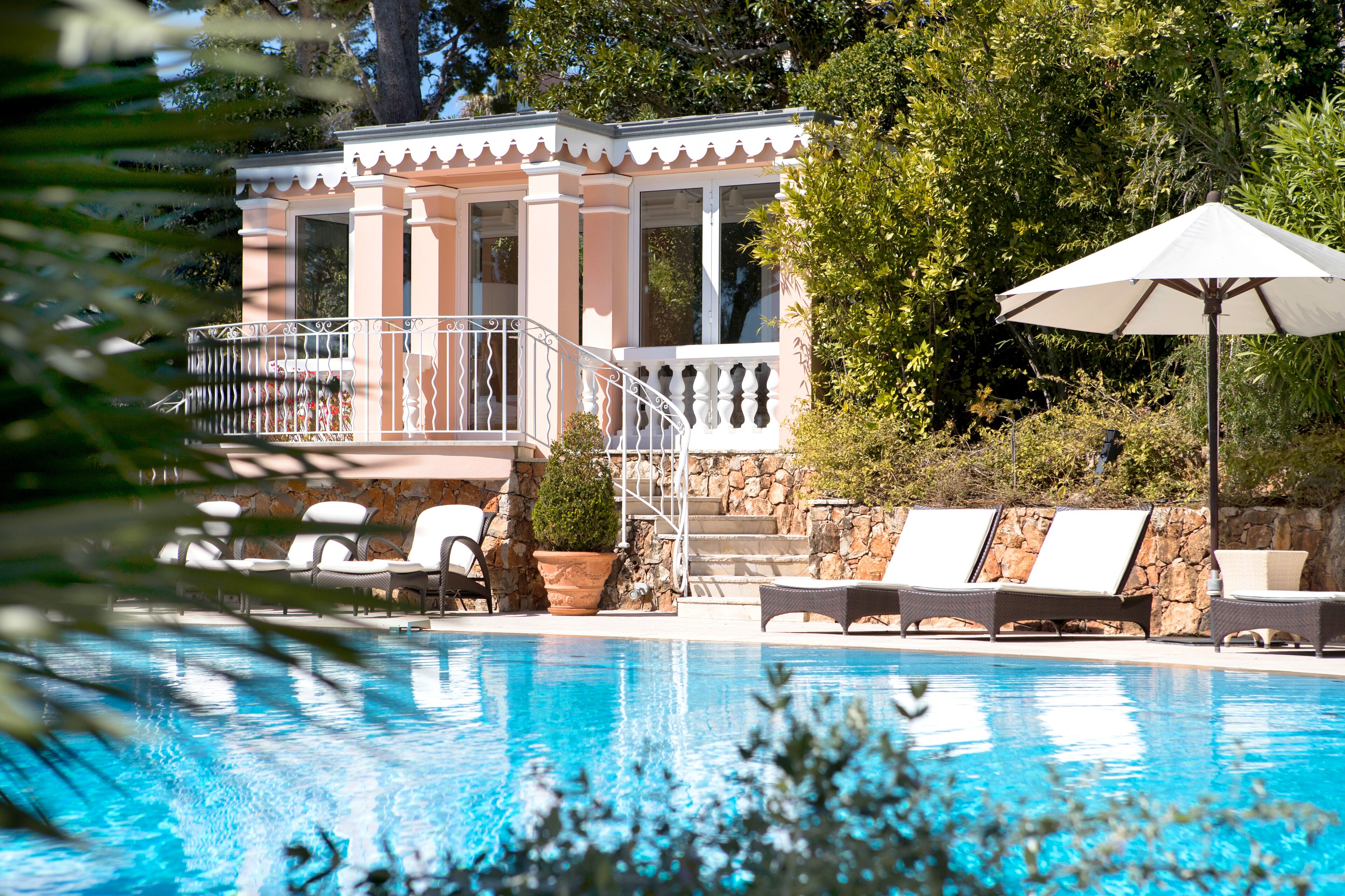 A pool area with lounge chairs and a covered umbrella, surrounded by a stone wall and lush greenery.
