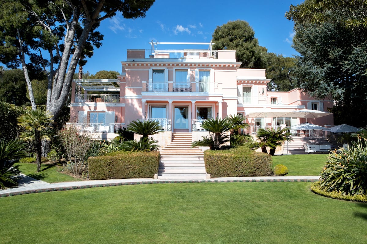 Villa rosa de dos pisos con entrada central, rodeada de jardines cuidados y palmeras.