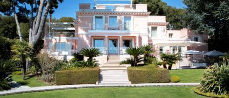 A two-story pink villa with a central entrance, surrounded by well-maintained gardens and palm trees.