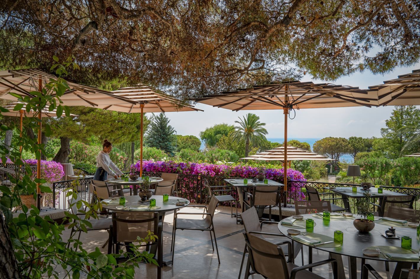 An outdoor dining area with tables, chairs, and umbrellas, surrounded by lush greenery and flowering plants.