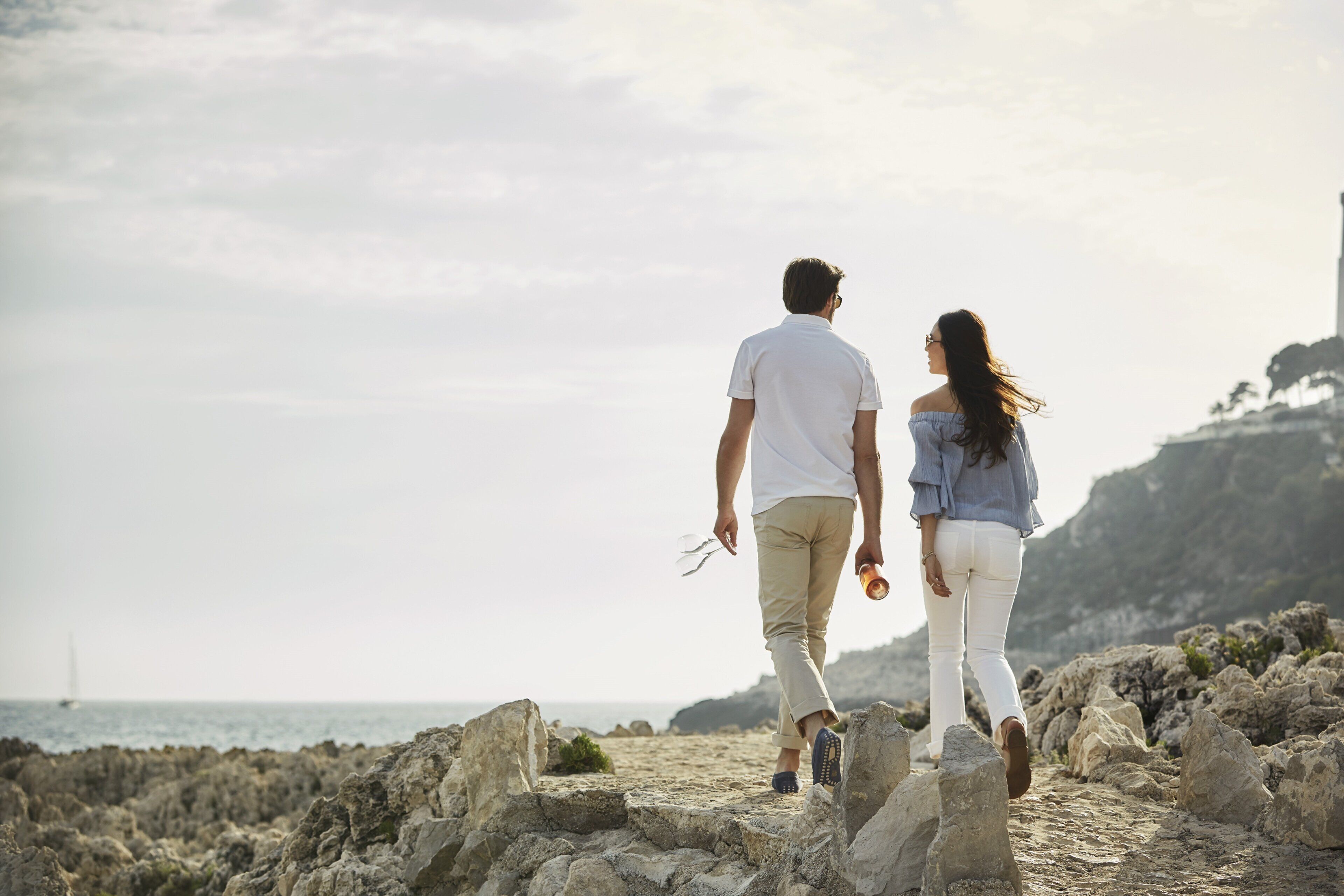 A couple walking on a rocky beach with a view of the sea and a distant sailboat.