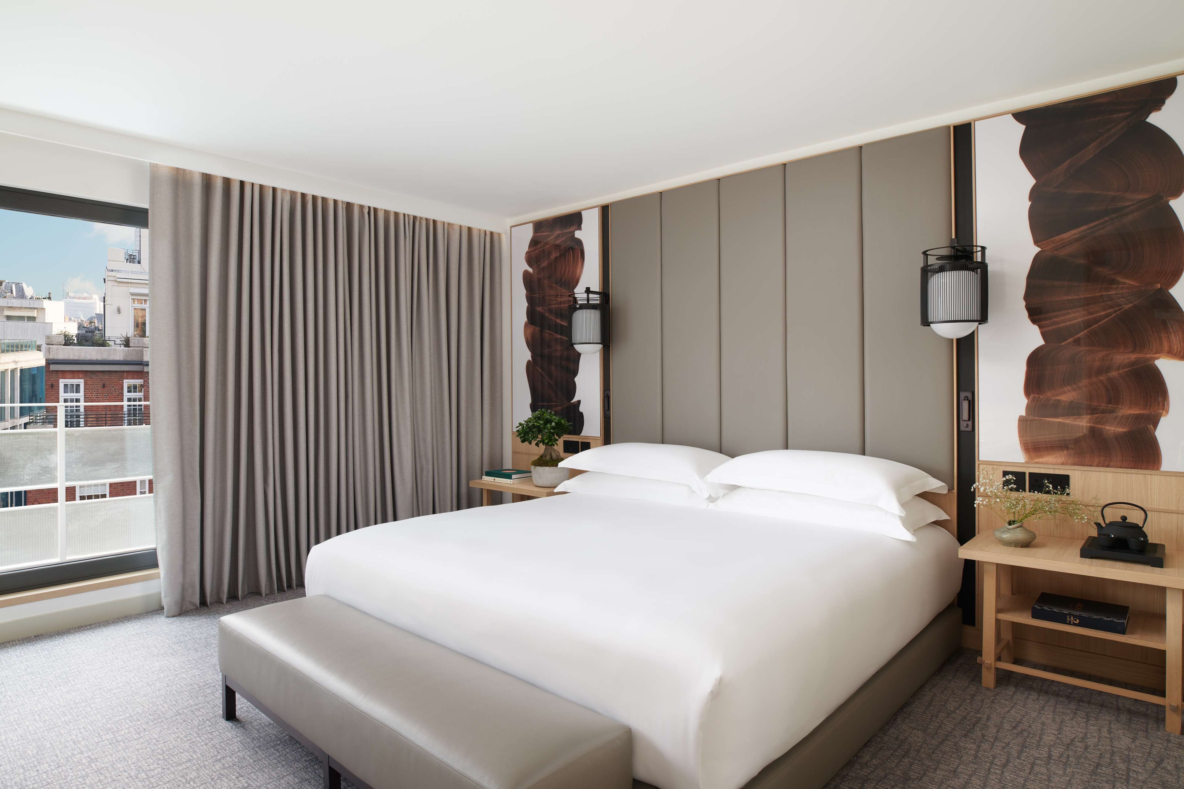 Een moderne hotelkamer met een groot bed, een bankje, een nachtkastje met een lamp, een vaas met bloemen en uitzicht op de stad door het raam.