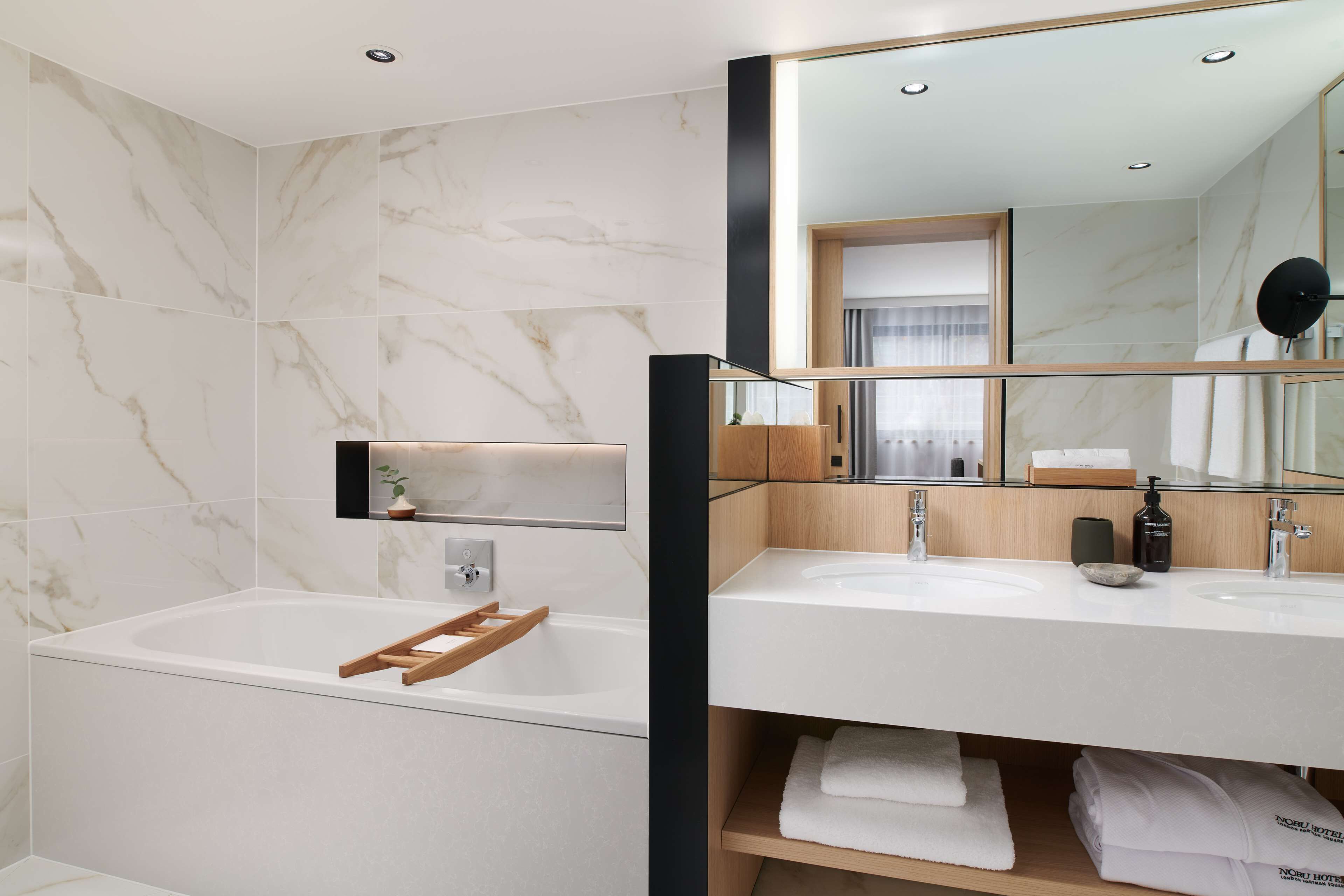 A modern bathroom with a marble wall, a wooden bath tray, a large mirror, and two sinks with a wooden shelf below.