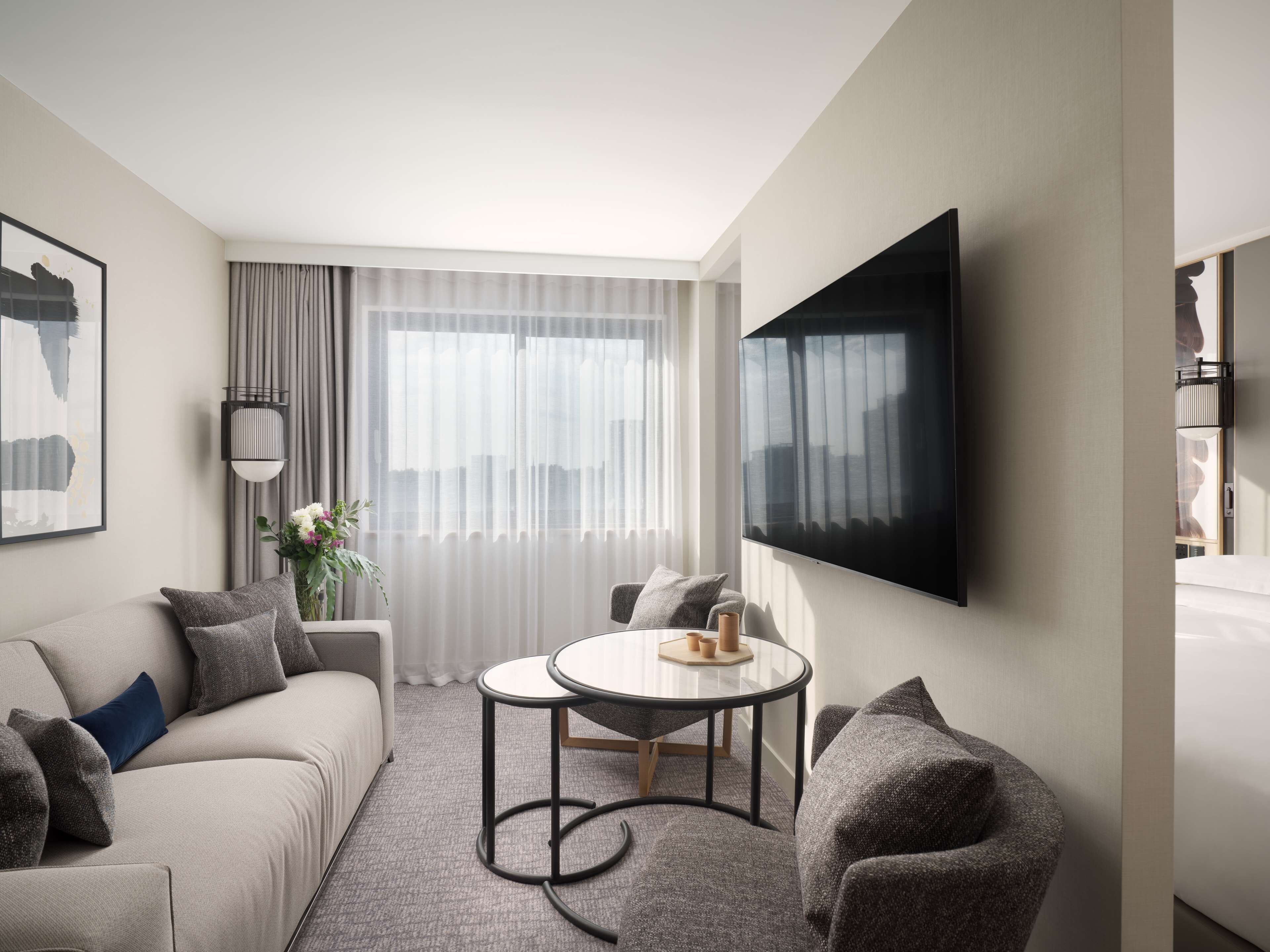 A modern hotel room with a sofa, armchair, round table, and a large TV on the wall.