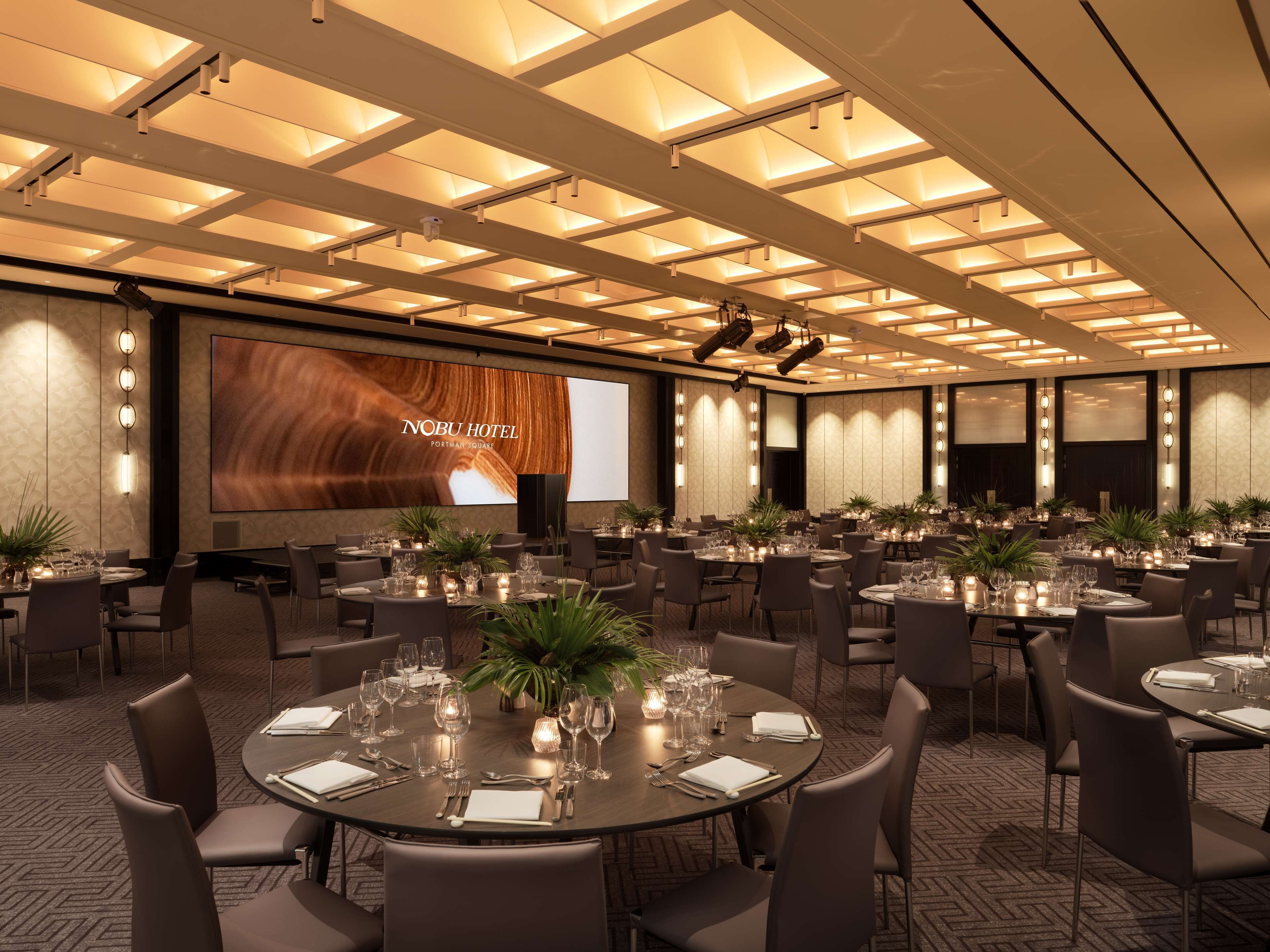 A conference room with multiple tables set for dining, a large screen displaying the hotel's name, and a modern ceiling design.