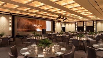 A conference room with multiple tables set for dining, a large screen displaying the hotel's name, and a modern ceiling design.