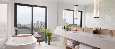 A modern bathroom with a large bathtub, a long vanity with a mirror, and a view of the city through large windows.