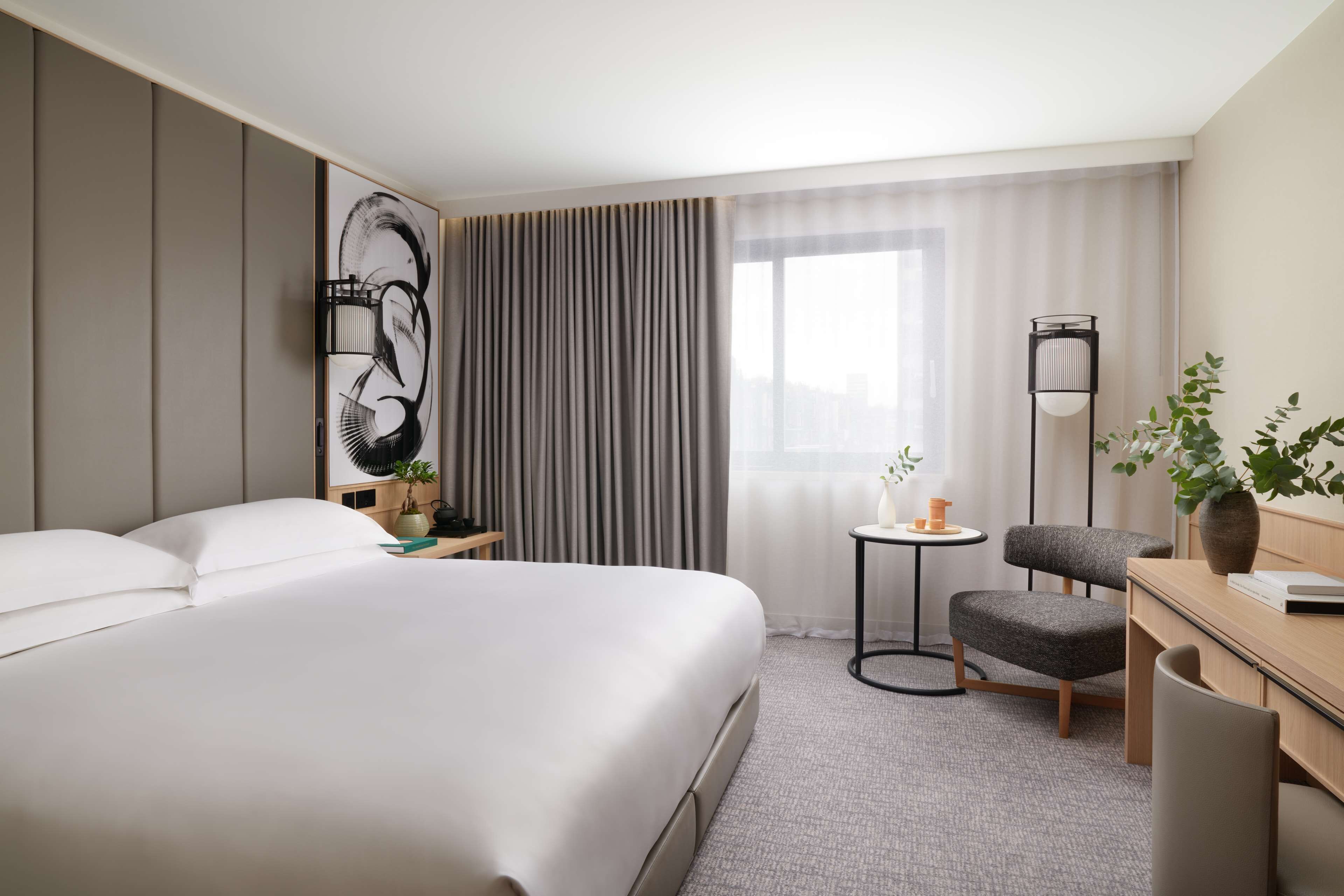 A hotel room with a large bed, a desk, a chair, a small table with a plant, and a large window with curtains.