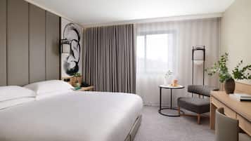 A hotel room with a large bed, a desk, a chair, a small table with a plant, and a large window with curtains.