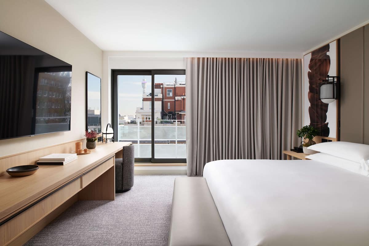 A modern hotel room with a large bed, a desk with a flat-screen TV, a view of the city through a large window, and a cityscape visible in the background.