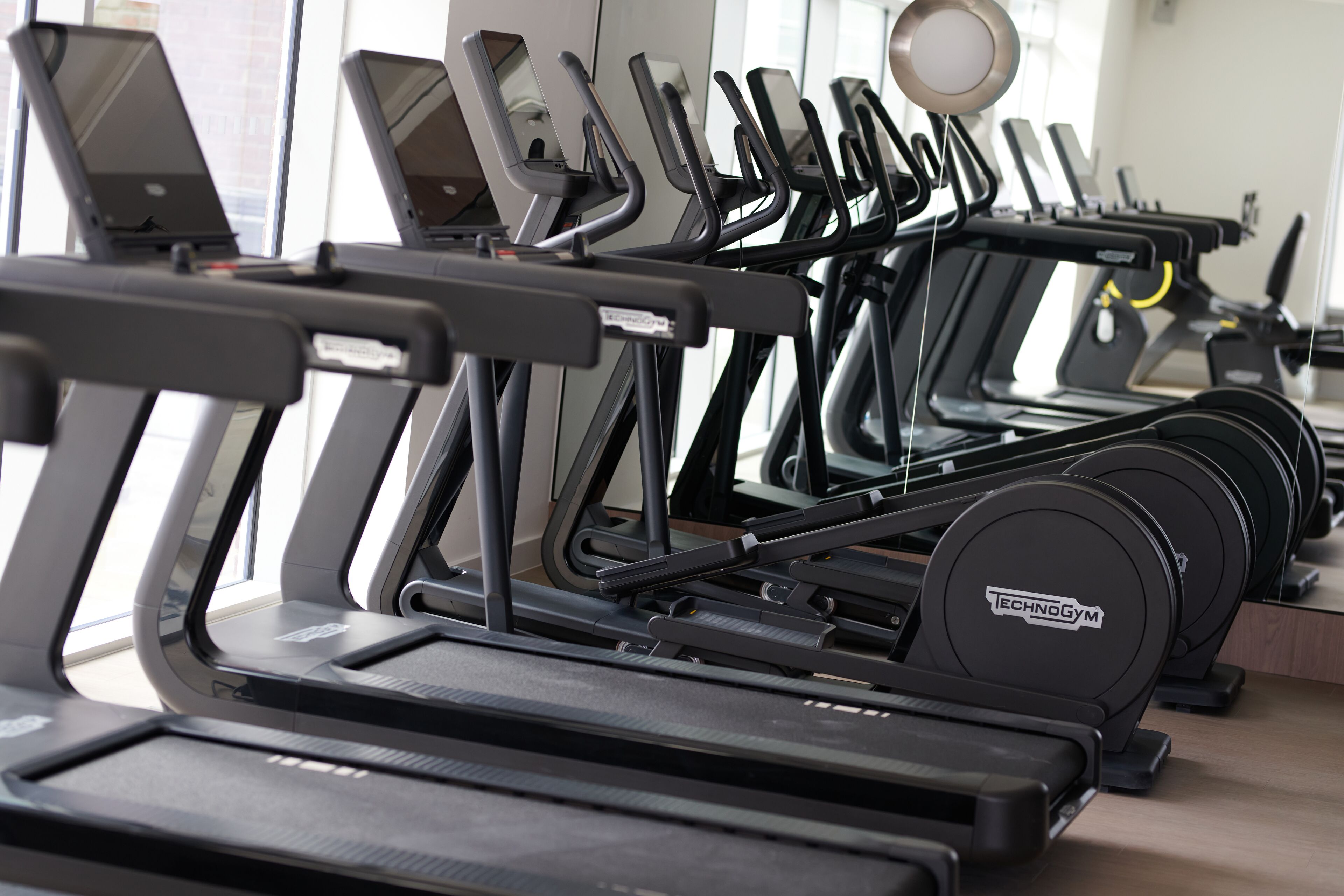 En række løbebånd med tilkoblede bærbare computere, der viser et fitnesscenter med integreret teknologi.