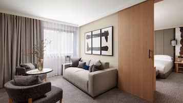 A modern hotel room with a sofa, armchair, and a small table with a vase. There is a bed with a wooden headboard and a nightstand with a lamp.