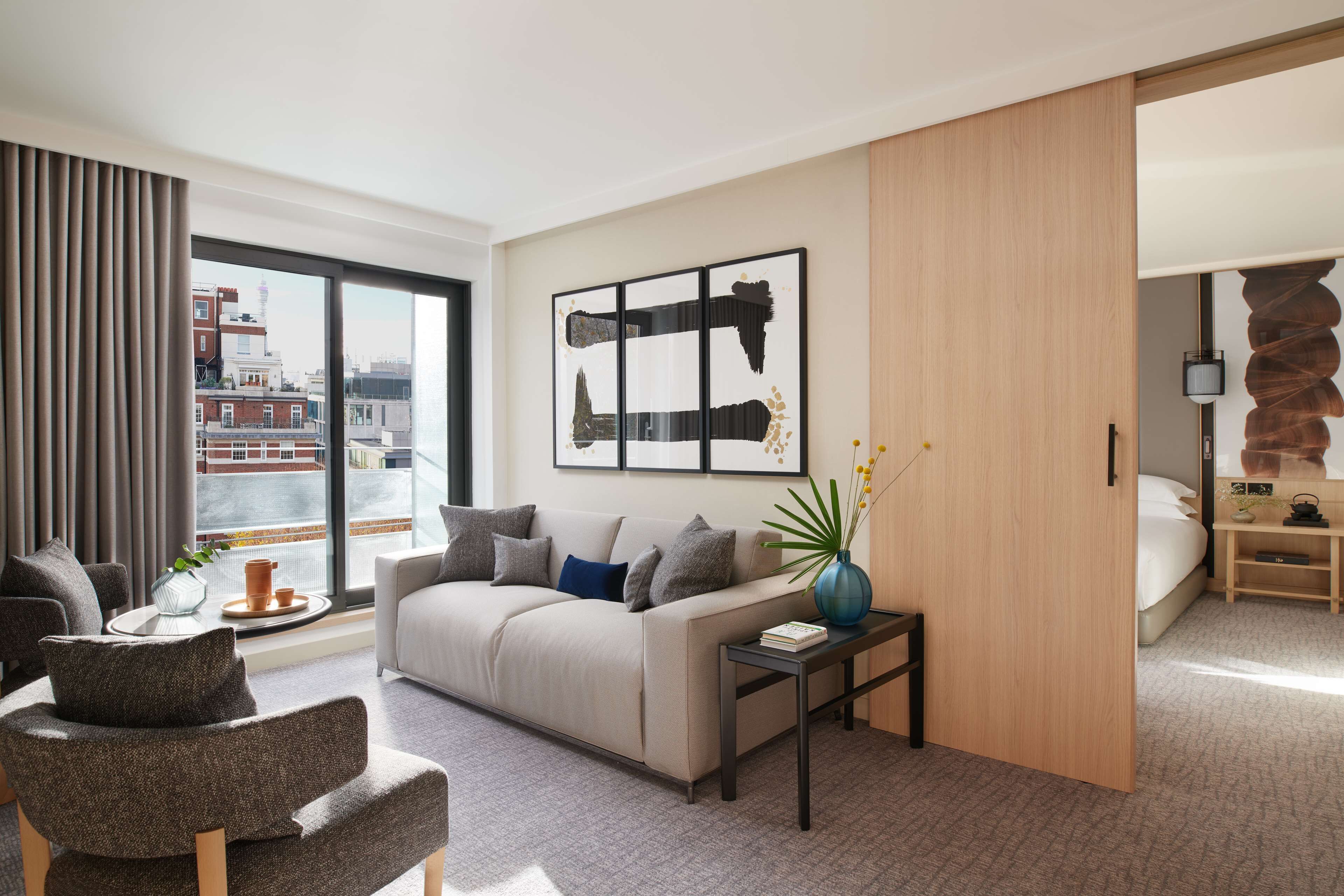 A modern living room with a sofa, armchair, and coffee table, a large window with a city view, and a piece of abstract art on the wall.
