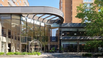A modern hotel with a curved glass facade, a prominent circular logo, and a well-maintained outdoor area with plants.