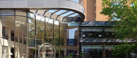 A modern hotel with a curved glass facade, a prominent circular logo, and a well-maintained outdoor area with plants.