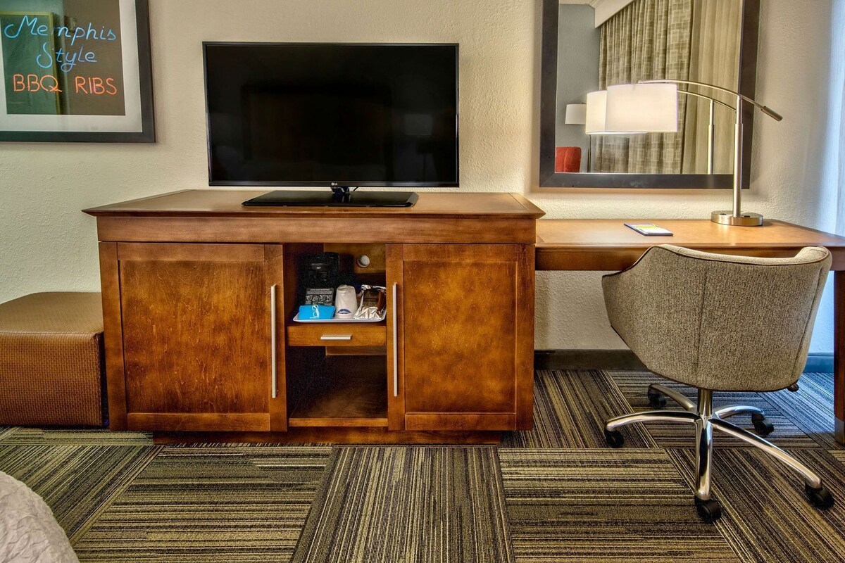 Une chambre d’hôtel avec un bureau en bois, une télévision, un miroir, une lampe, une chaise et un petit coin cuisine.