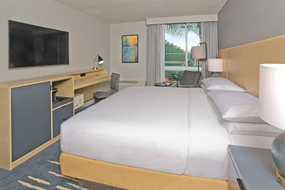 A hotel room with a large bed, a desk with a chair, a TV, a painting, and a window with curtains.