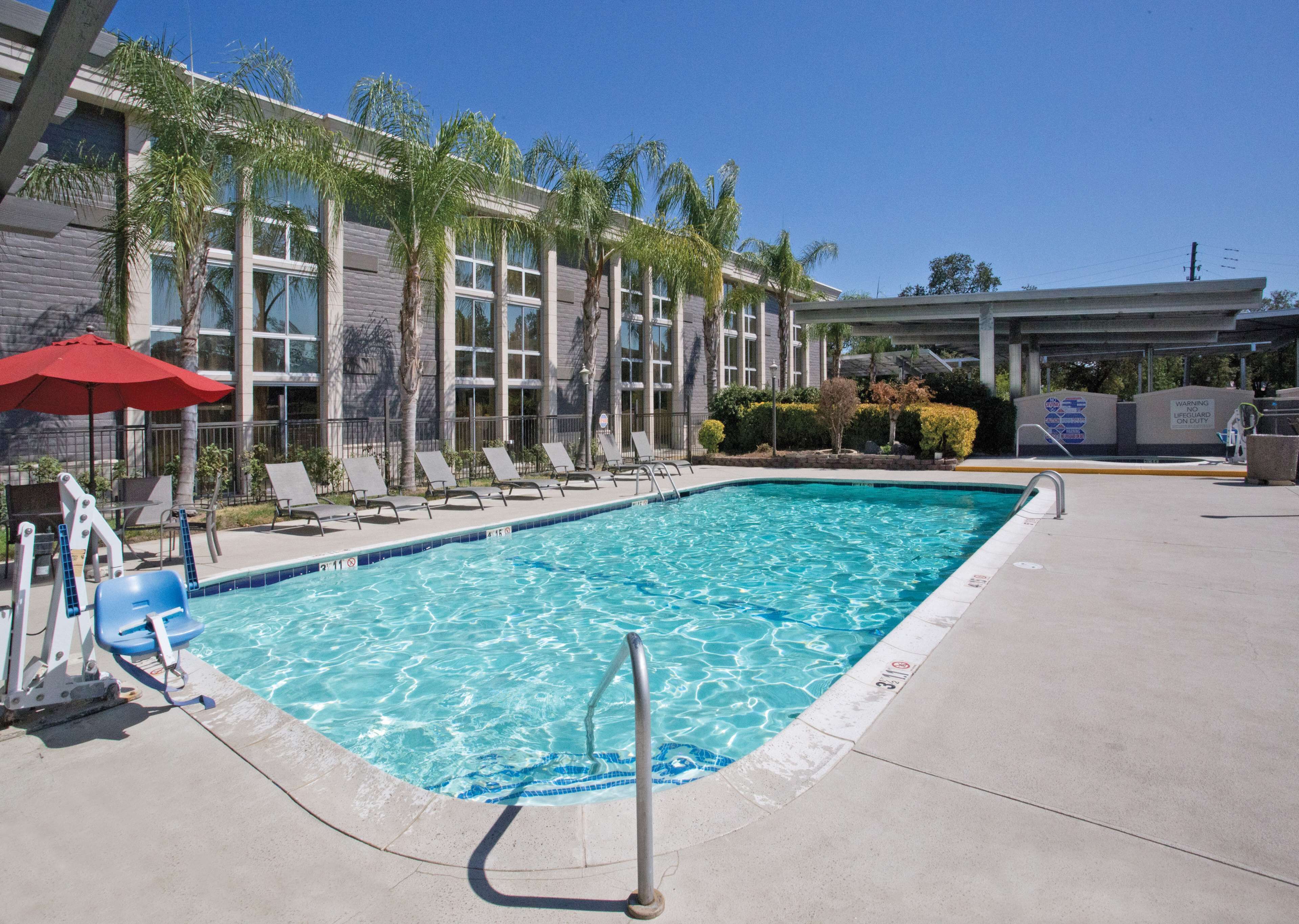 A swimming pool with lounge chairs and a red umbrella, surrounded by palm trees and a building with large windows.