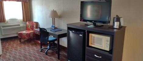 Habitación de hotel con escritorio, televisor, microondas, refrigerador, silla, sofá y ventana con cortinas.