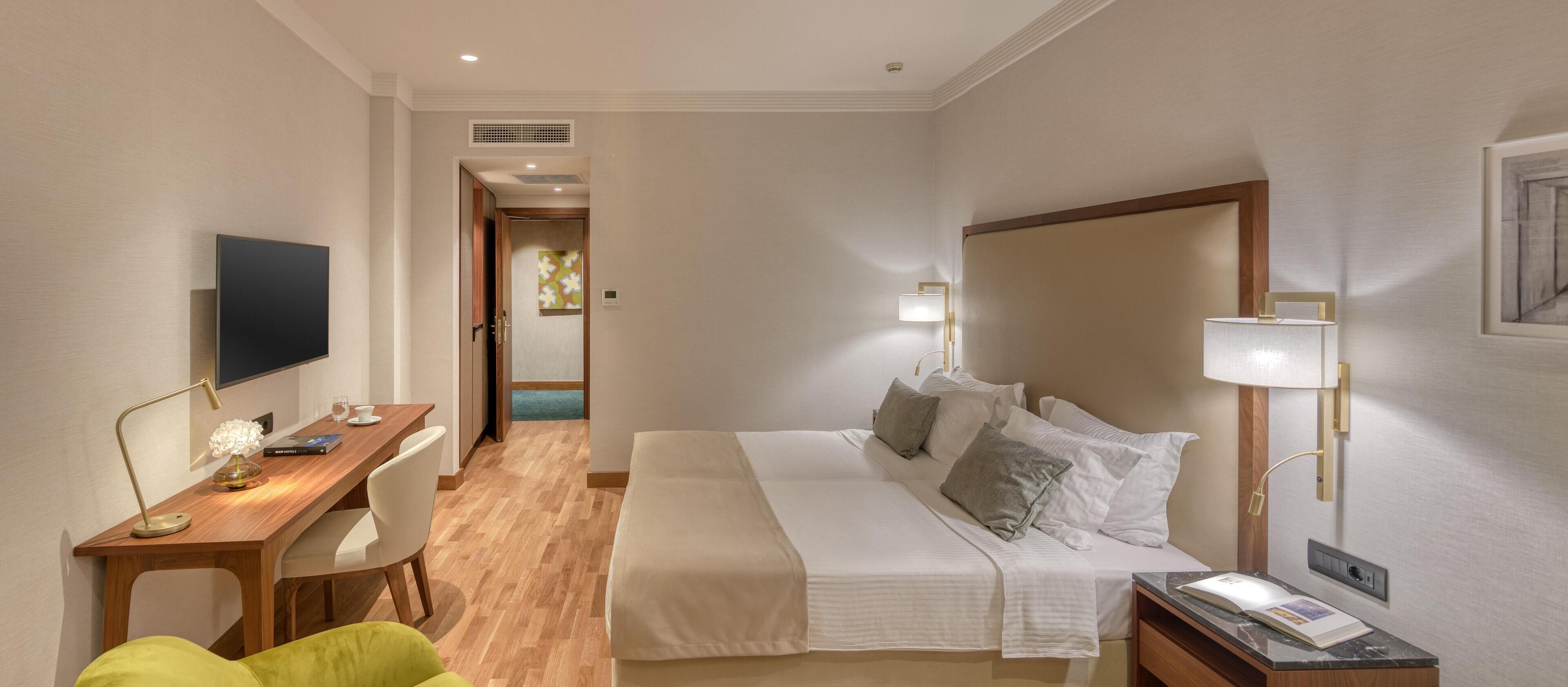 Une chambre d’hôtel comprenant un lit, une table de chevet, une lampe, une télévision, un bureau, une chaise et donnant sur le couloir.