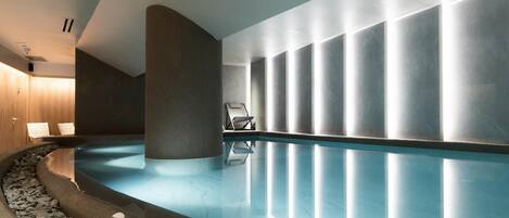 Una piscina interior moderna con superficie de agua cristalina, borde de piedra y diseño minimalista, con un pilar grande y una fila de luces a lo largo de la pared.