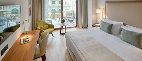 A hotel room with a bed, a desk, a chair, a small table with a plant, and a balcony with a view of buildings.