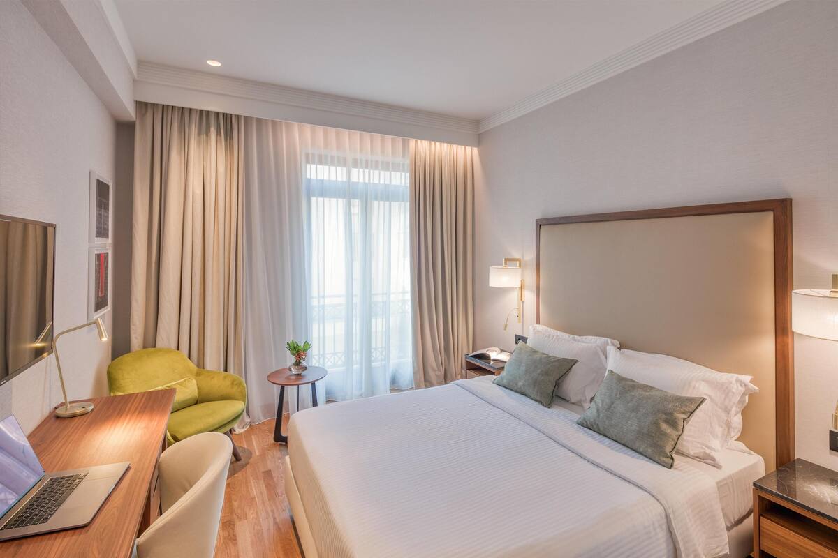 Une chambre d’hôtel avec un grand lit, un bureau avec un ordinateur portable, un fauteuil vert et une petite table avec une plante.