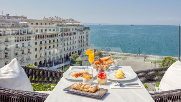 A table with breakfast items and a glass of orange juice, overlooking a coastal cityscape.