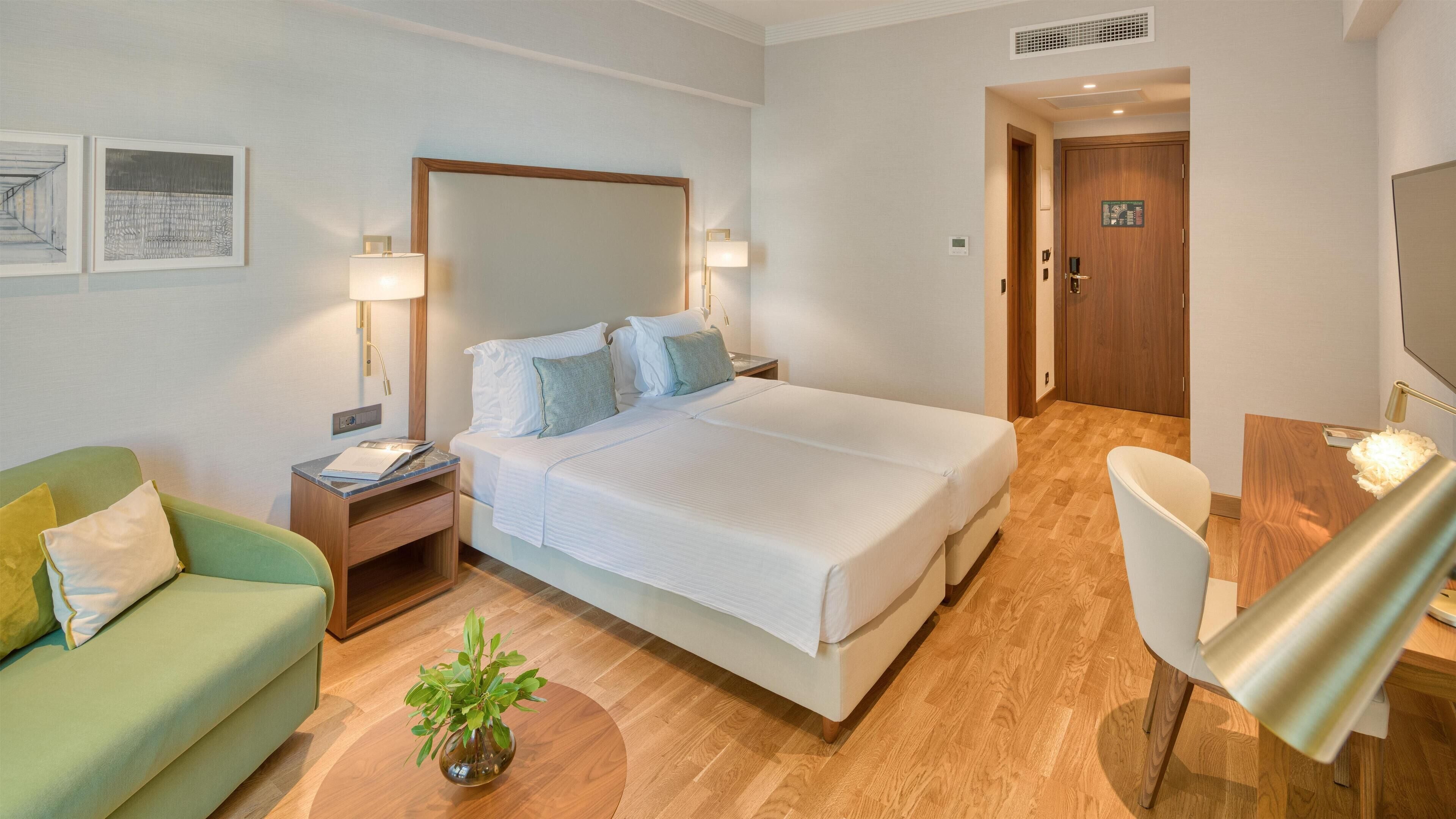 A hotel room with a large bed, a desk, a chair, a nightstand, a lamp, and a plant.