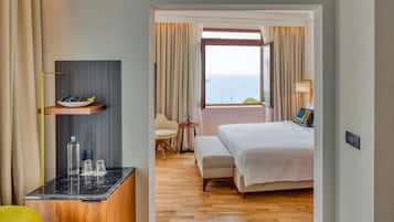 A hotel room with a bed, a small desk with a bowl of fruit, a chair, a window with a view of the sea, and a wooden floor.