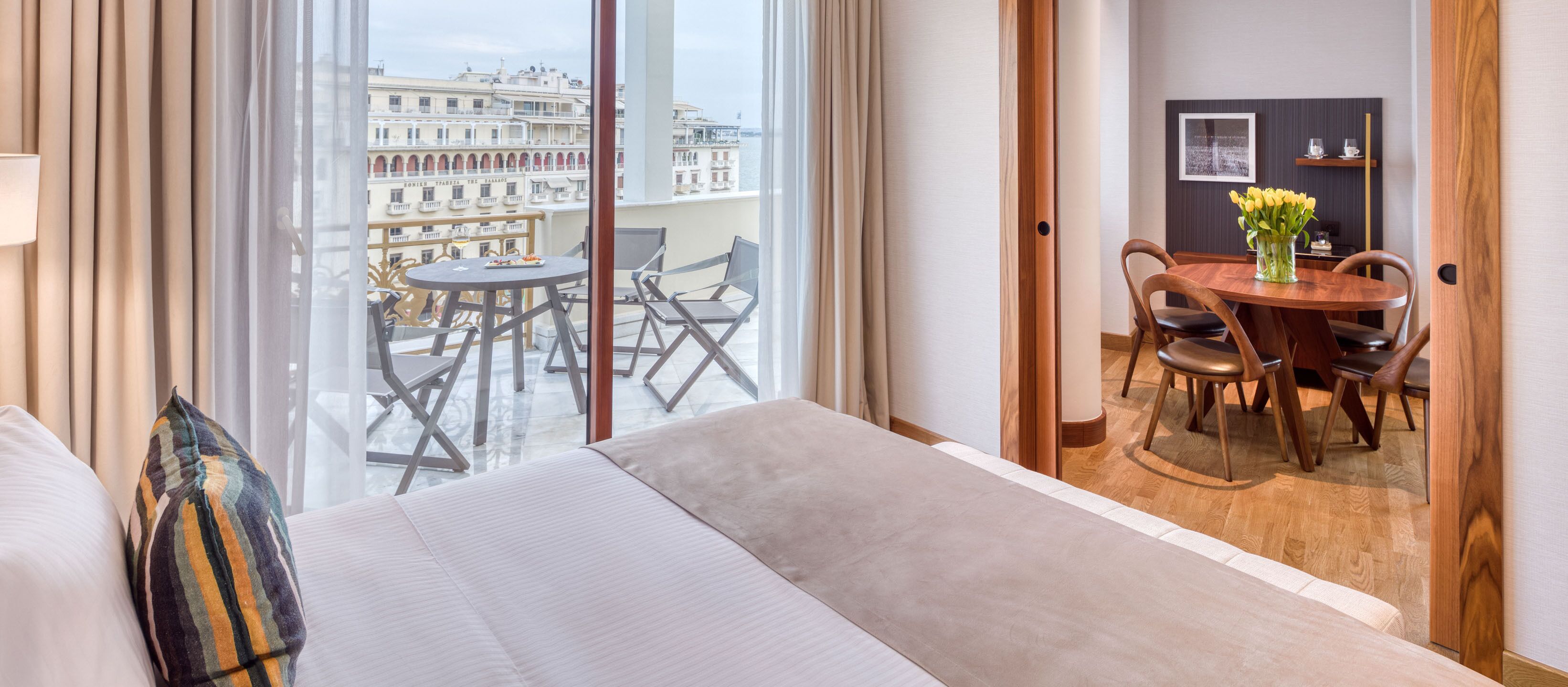 A hotel room with a large bed, a balcony with a dining table and chairs, and a view of the cityscape.