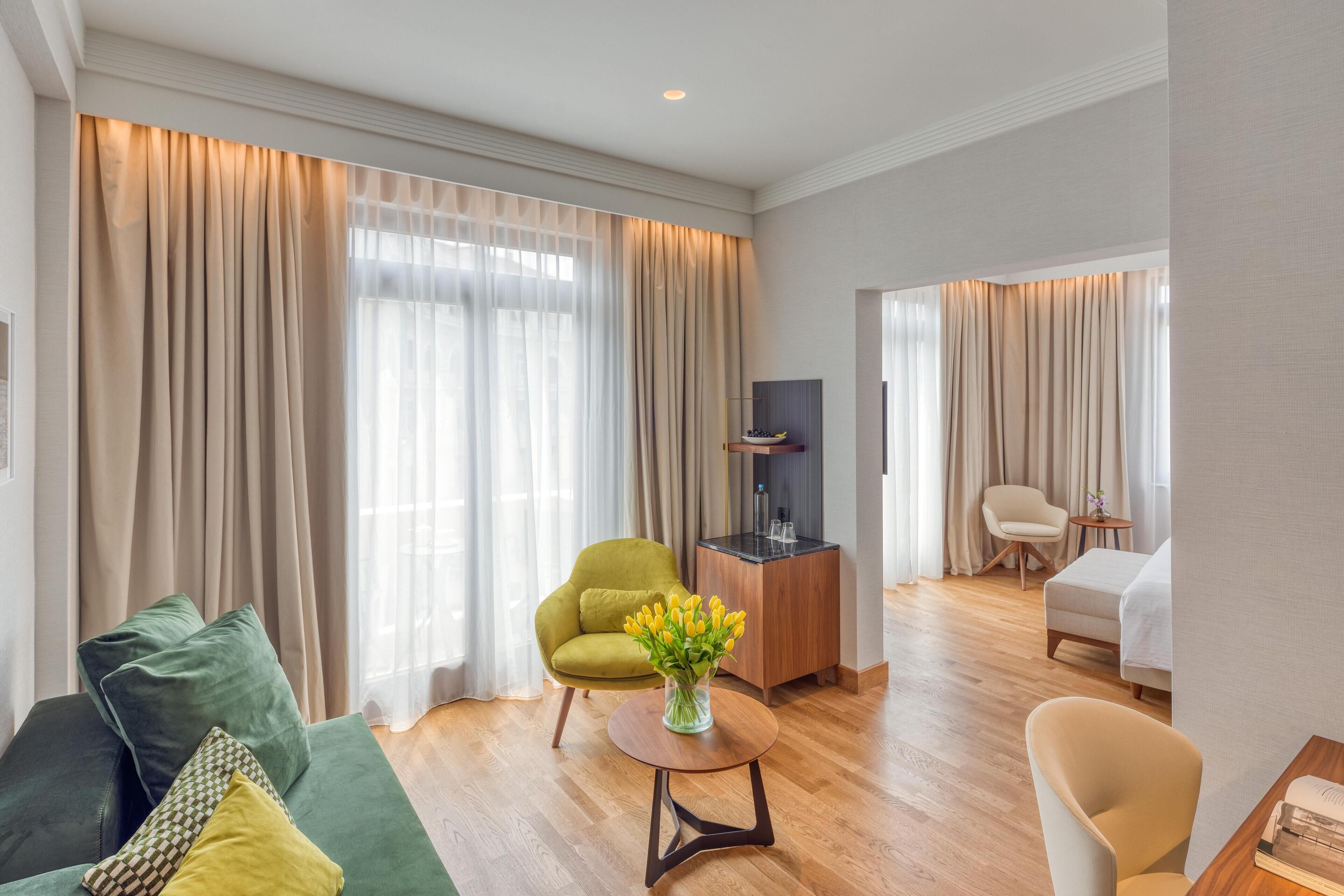 A modern hotel room with a green sofa, a round wooden table with a vase of flowers, a small wooden side table, a bed, a TV on a wooden stand, and a chair.