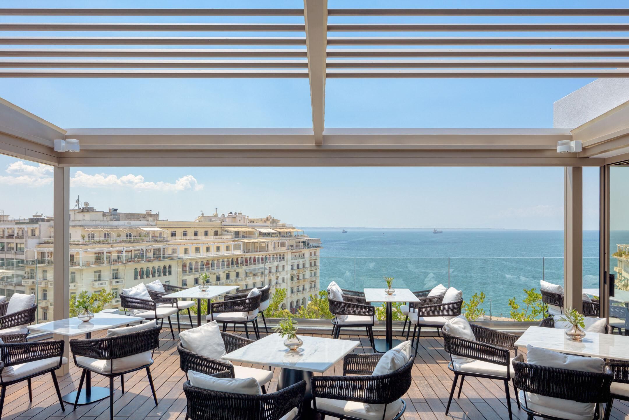 A terrace with outdoor seating, a view of the sea, and a cityscape in the background.