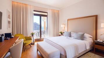 A hotel room with a large bed, a desk with a chair, a small table with a plant, and a view of the city through the window.