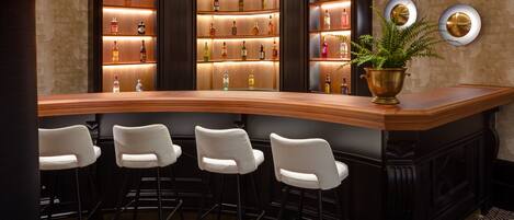 Zona de bar con una barra curva, tres taburetes blancos y un mueble de madera con estantes iluminados que exhiben botellas.